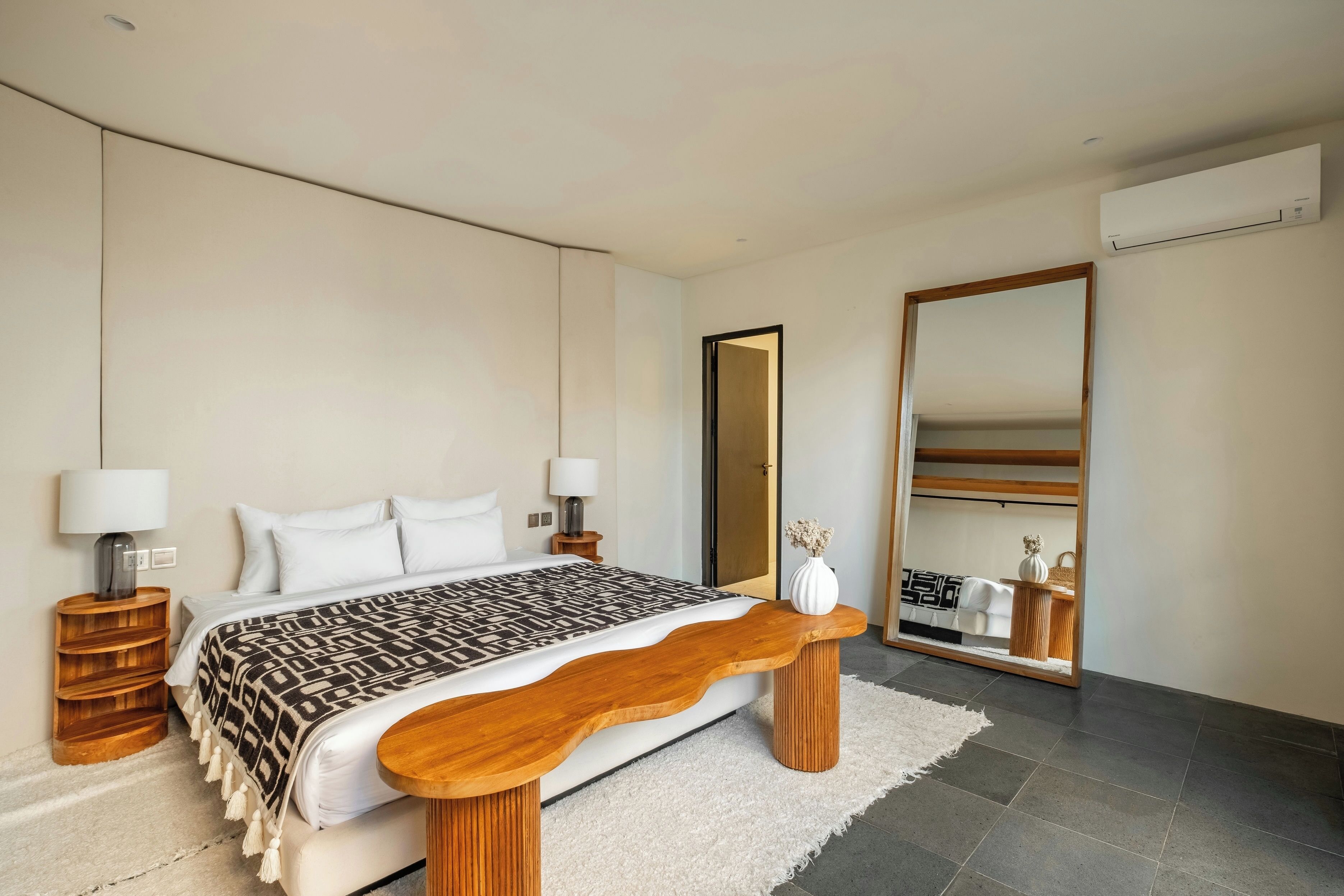 A modern bedroom with a large bed, bedside lamps, a wooden bench, a mirror, and a wardrobe.