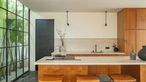 A modern kitchen with a large island, wooden stools, and a view of green plants through a glass partition. - Casa Siella (Kerobokan)