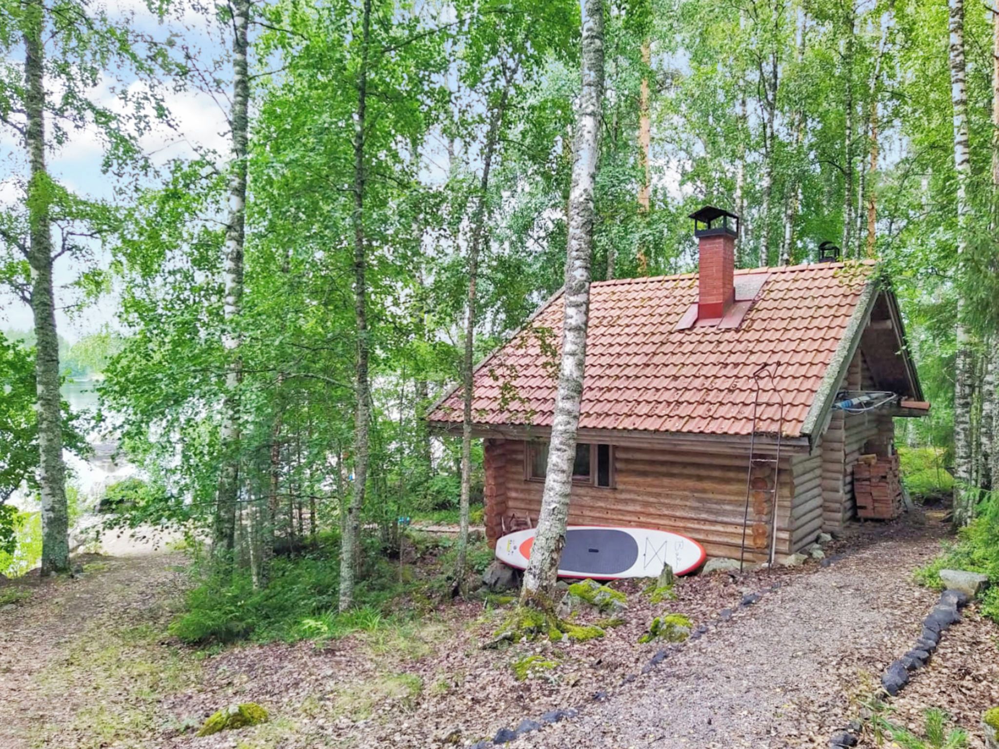 En stuga av trä med ett tak täckt av tegelpannor, omgiven av träd, och en paddelbräda lutad mot väggen.