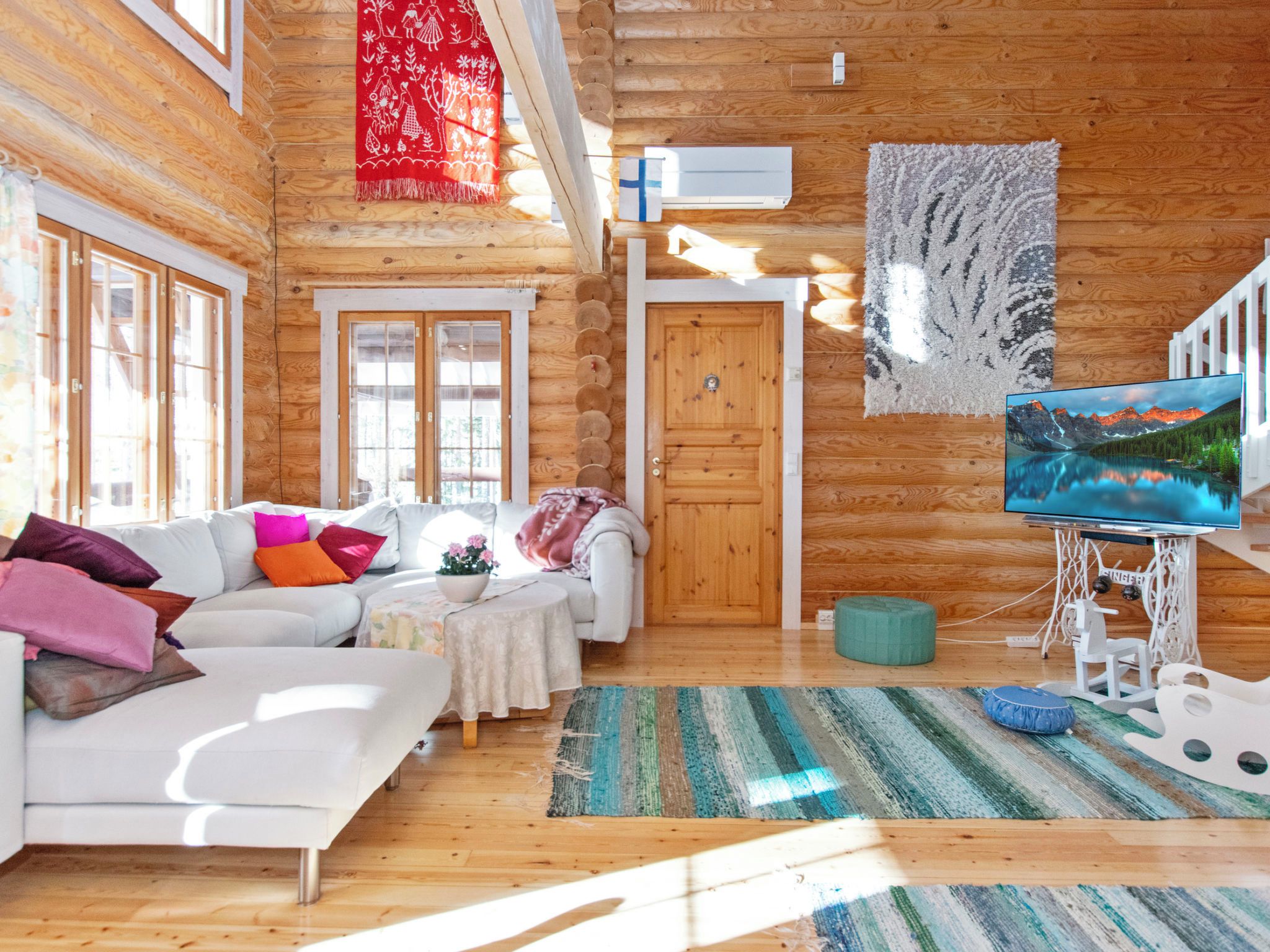 L’intérieur d’un chalet confortable, avec des murs en bois, un canapé blanc, des coussins colorés, un téléviseur et une grande fenêtre.