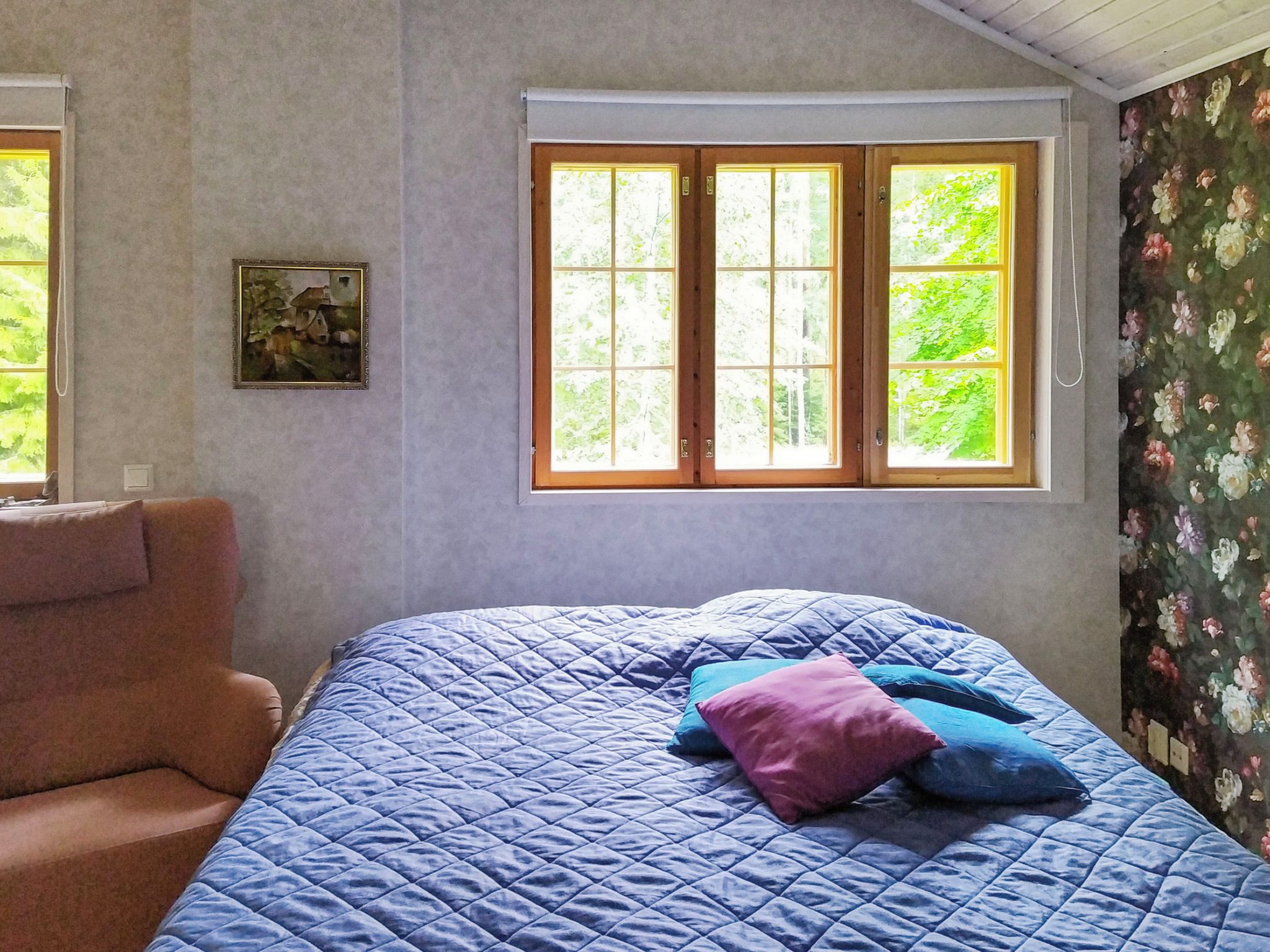 Ett sovrum med en säng, en stol, ett fönster med utsikt över grönområden och en tavla på väggen.