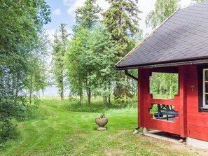 Une cabane rouge avec un jardinière en pierre devant, entourée d’une zone herbeuse et d’arbres.