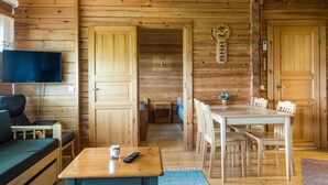 Interior de una cabaña de madera con televisión, sofá, mesa de comedor, sillas y una mesita con control remoto.