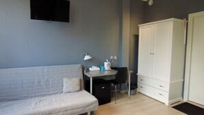 Een kamer met een witte bank, een bureau met lamp, een kleine koelkast, een witte kledingkast en een televisie aan de muur.