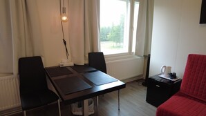 Une pièce comprenant une table, des chaises, un radiateur, une fenêtre avec des rideaux, une chaise rouge et une petite table sur laquelle se trouvent une bouilloire et des tasses.