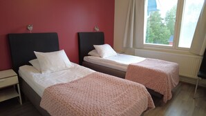 Une chambre avec deux lits, chacun étant équipé de draps blancs et d’une couverture rose. Une fenêtre offre une vue sur les arbres et un bâtiment à l’extérieur.