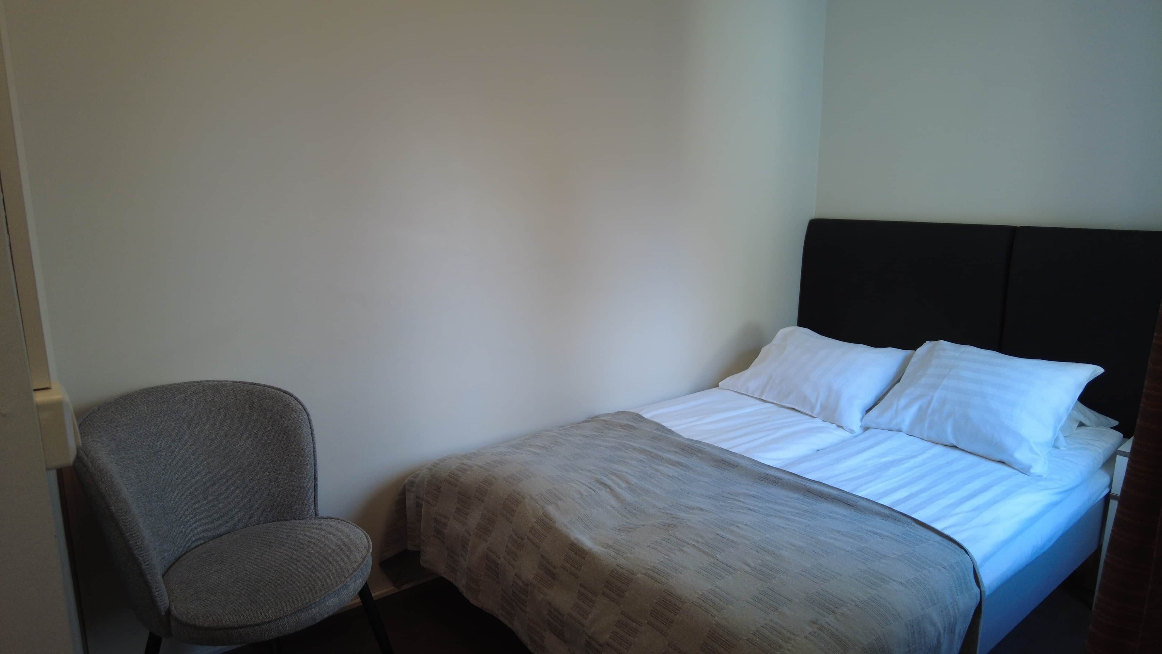 Un lit simple avec du linge de lit blanc, un fauteuil recouvert d’un tissu gris et un mur uni.