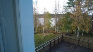 Une terrasse en bois avec une rambarde en métal, donnant sur une zone herbeuse parsemée d’arbres, avec un plan d’eau en arrière-plan.