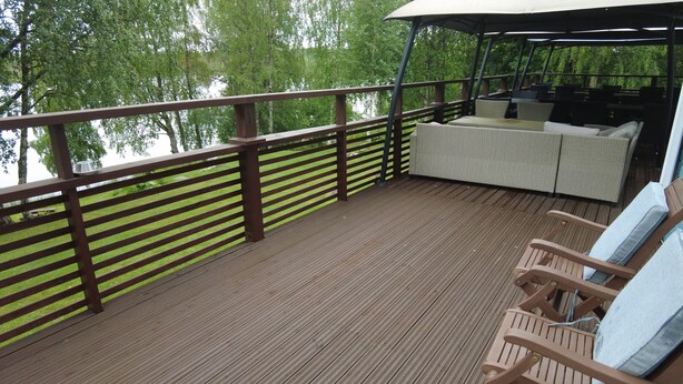 Una terraza de madera con barandilla, una zona de estar cubierta con cojines y vistas a un lago.