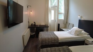 Una habitación de hotel pequeña y bien iluminada, con una cama, un escritorio con silla, un televisor montado en la pared y una ventana con cortinas.