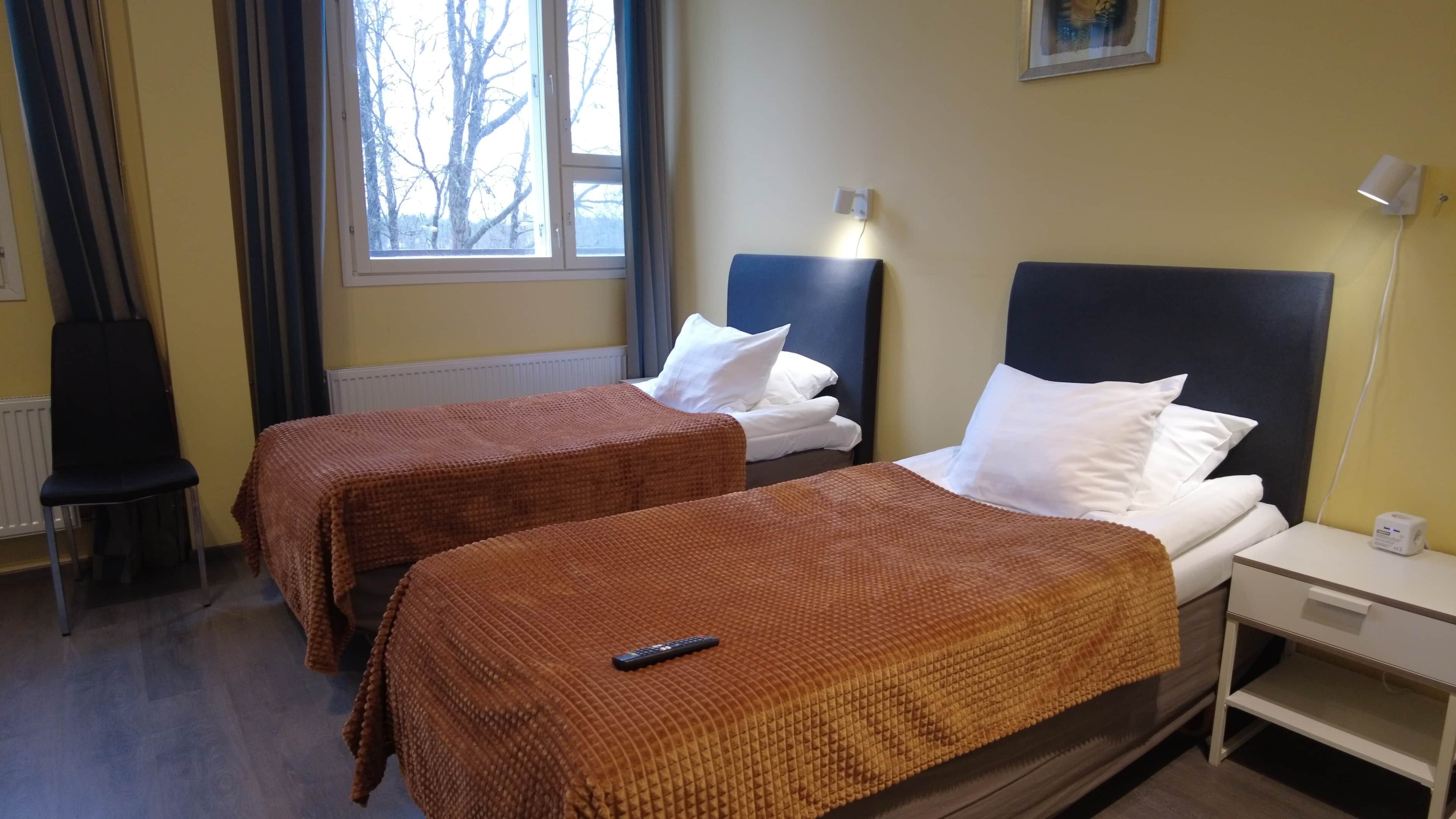 A hotel room with two beds, a chair, a nightstand, and a window with a view of trees.