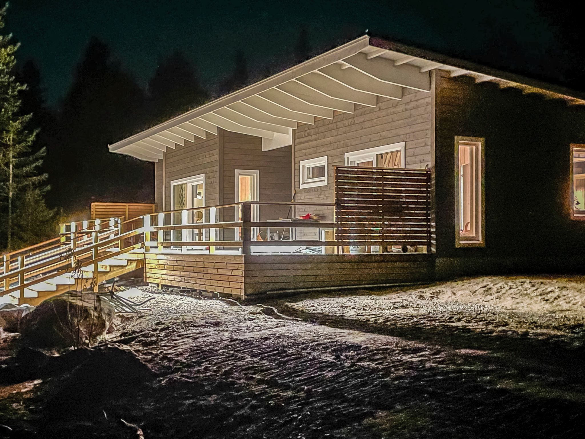 A two-story wooden cabin with a covered porch and a small deck, surrounded by a forested area.