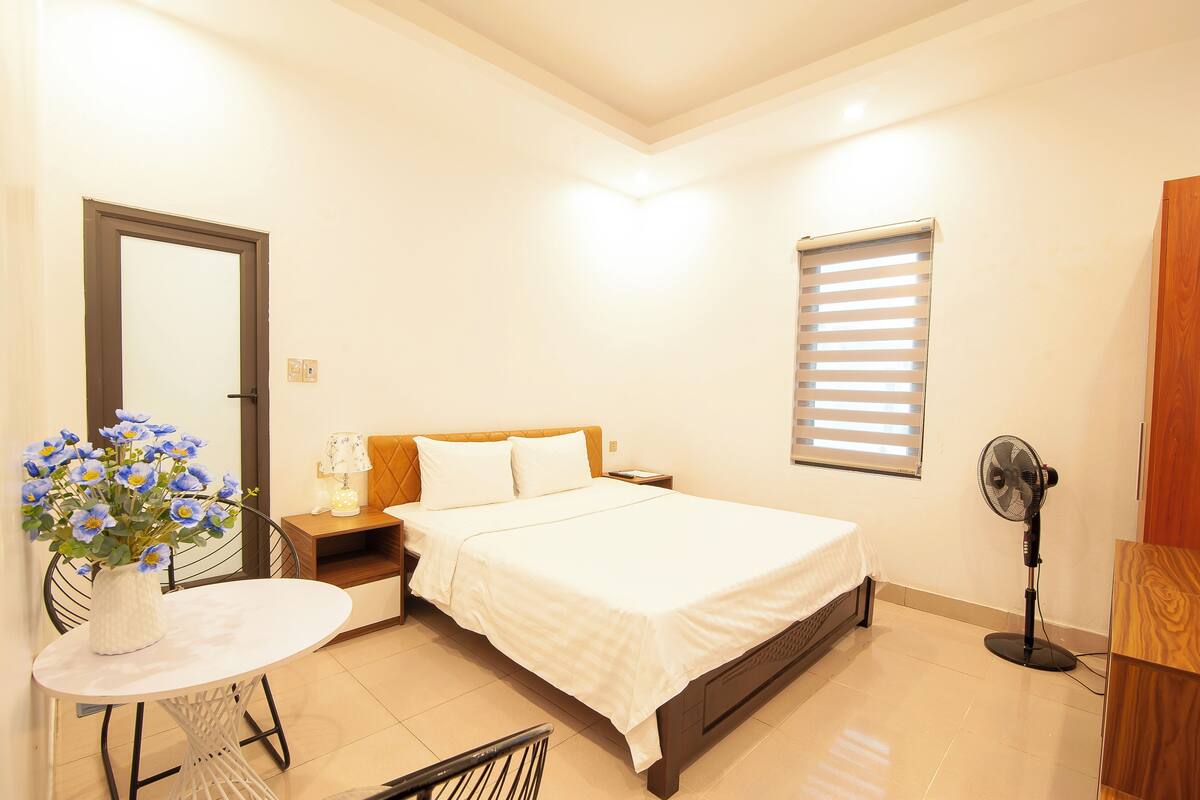 A bedroom with a bed, bedside table, lamp, and a standing fan.