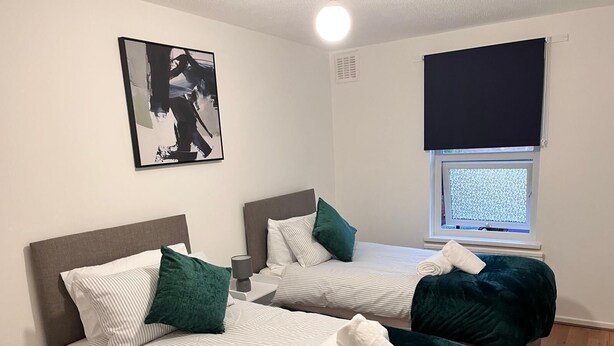 A room with two beds, each with a headboard, a bedside table, and a lamp. There is a window with a view of trees and a dark blind covering the window.