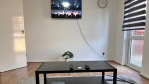 A room with a black TV on the wall, a clock, a plant, and a coffee table with remotes and a magazine.
