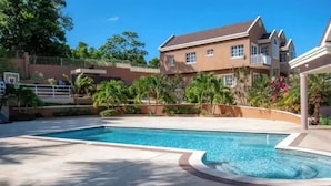 A large swimming pool with a curved edge, surrounded by a landscaped area with palm trees and a large two-story building in the background. - Luxury Sea-View Townhouse, Pool , Ocho Rios (Ocho Rios)