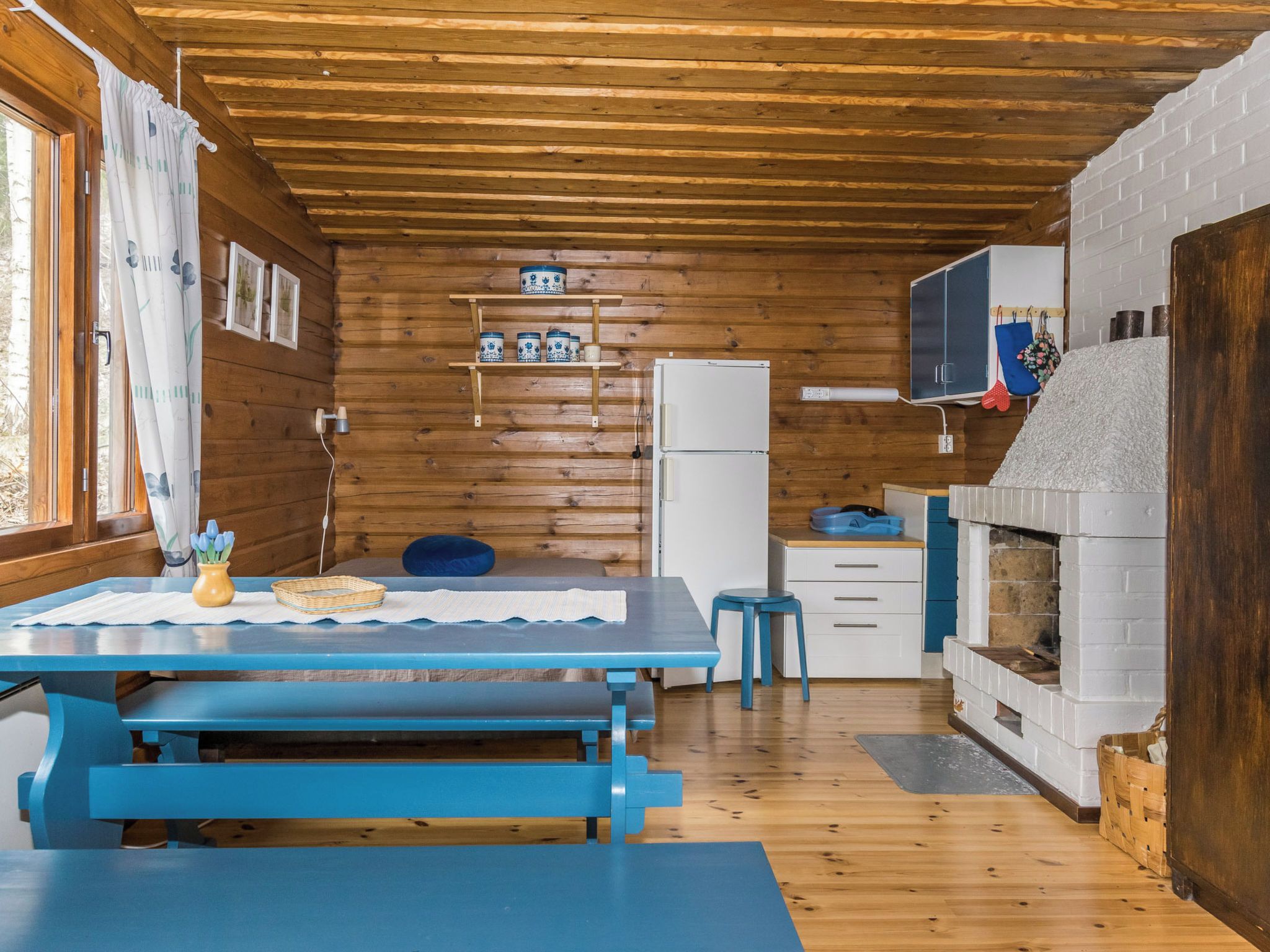 Interior d'una cabana de fusta amb taula de menjador, seients, nevera, prestatgeria amb objectes decoratius i una llar de foc.