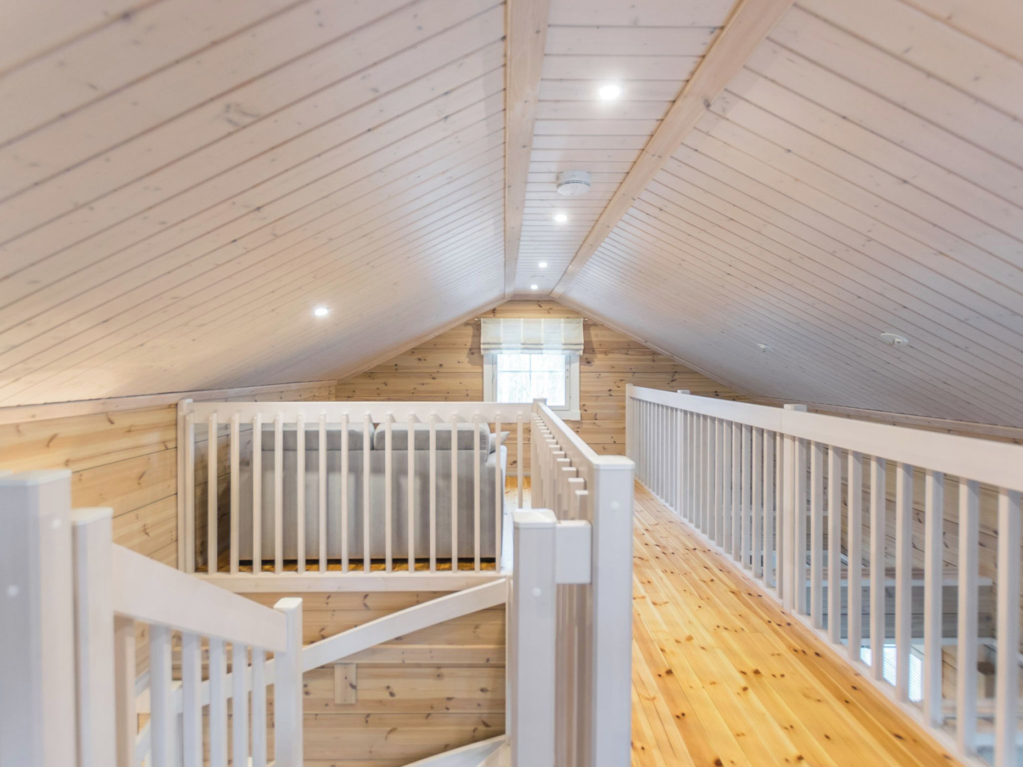 Una stanza mansardata con soffitto alto, travi in legno, ringhiera bianca e una finestra.