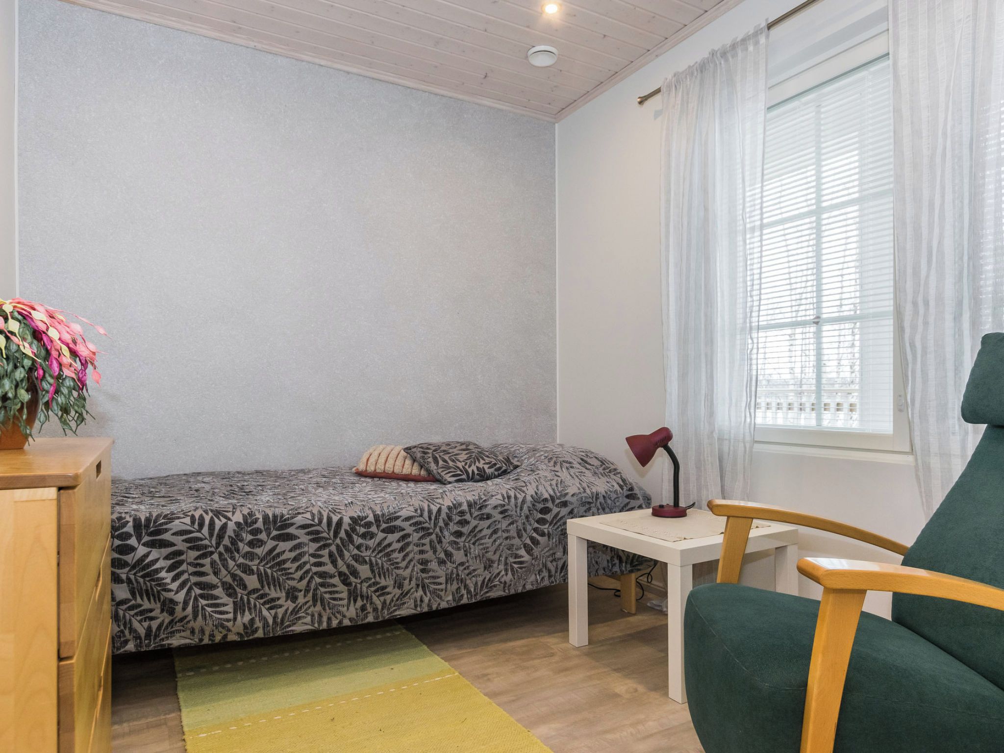 A small, neatly arranged room with a bed, a desk, a chair, and a window with curtains.