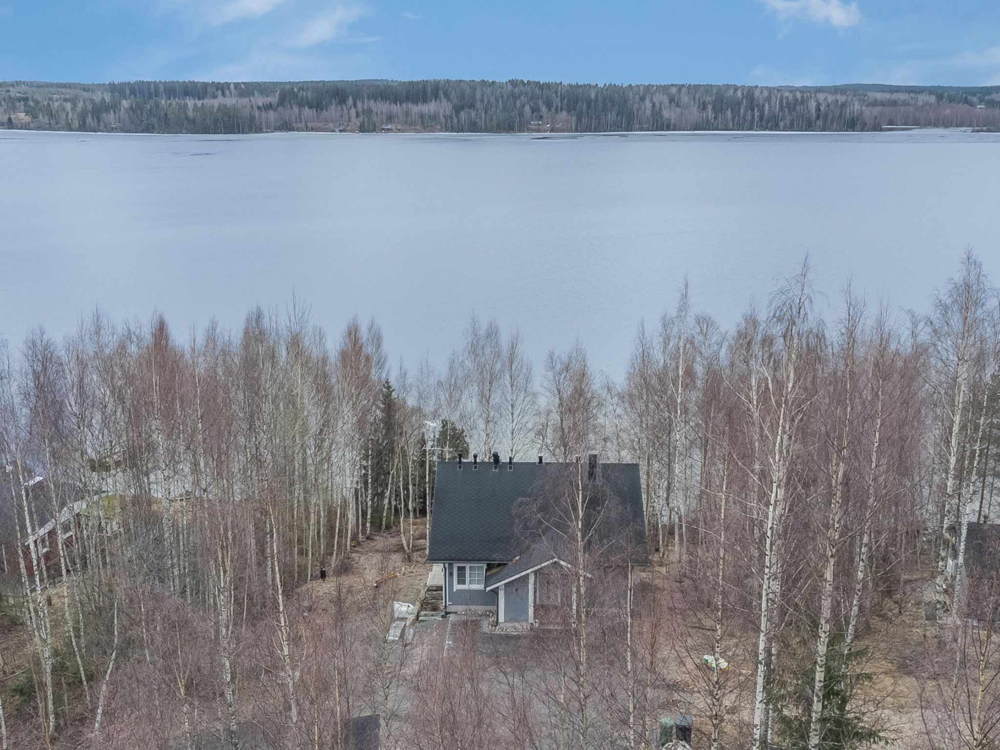 A house with a dark roof, surrounded by leafless trees, near a large body of water.