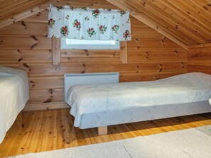 A small, cozy room with wooden walls and a sloped ceiling, featuring two single beds and a window with a floral curtain.