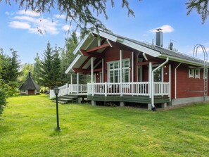 A red cabin with a white porch and a chimney, surrounded by a grassy yard and trees.