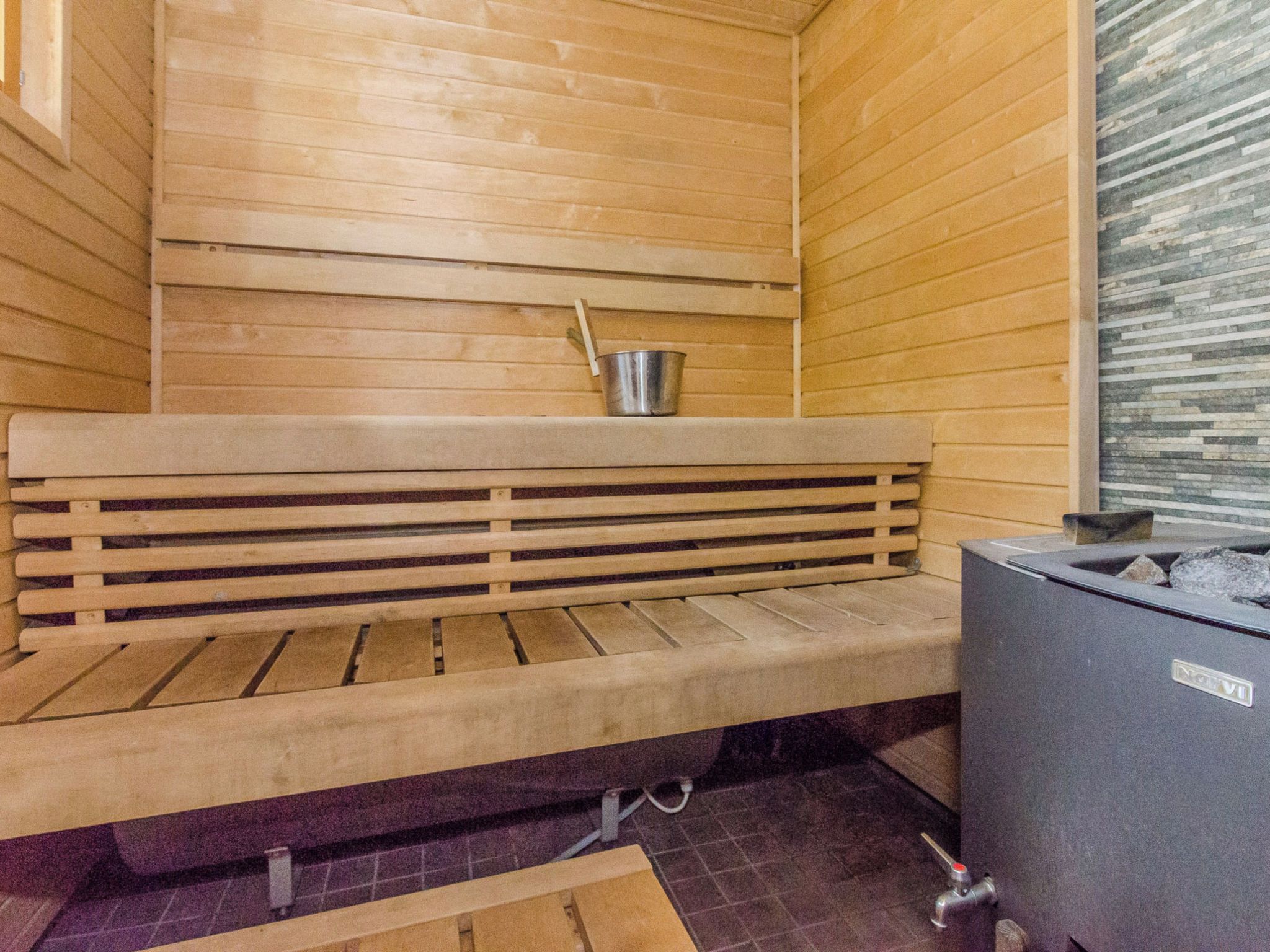 A sauna with a wooden bench, a metal bucket, and a stone wall.