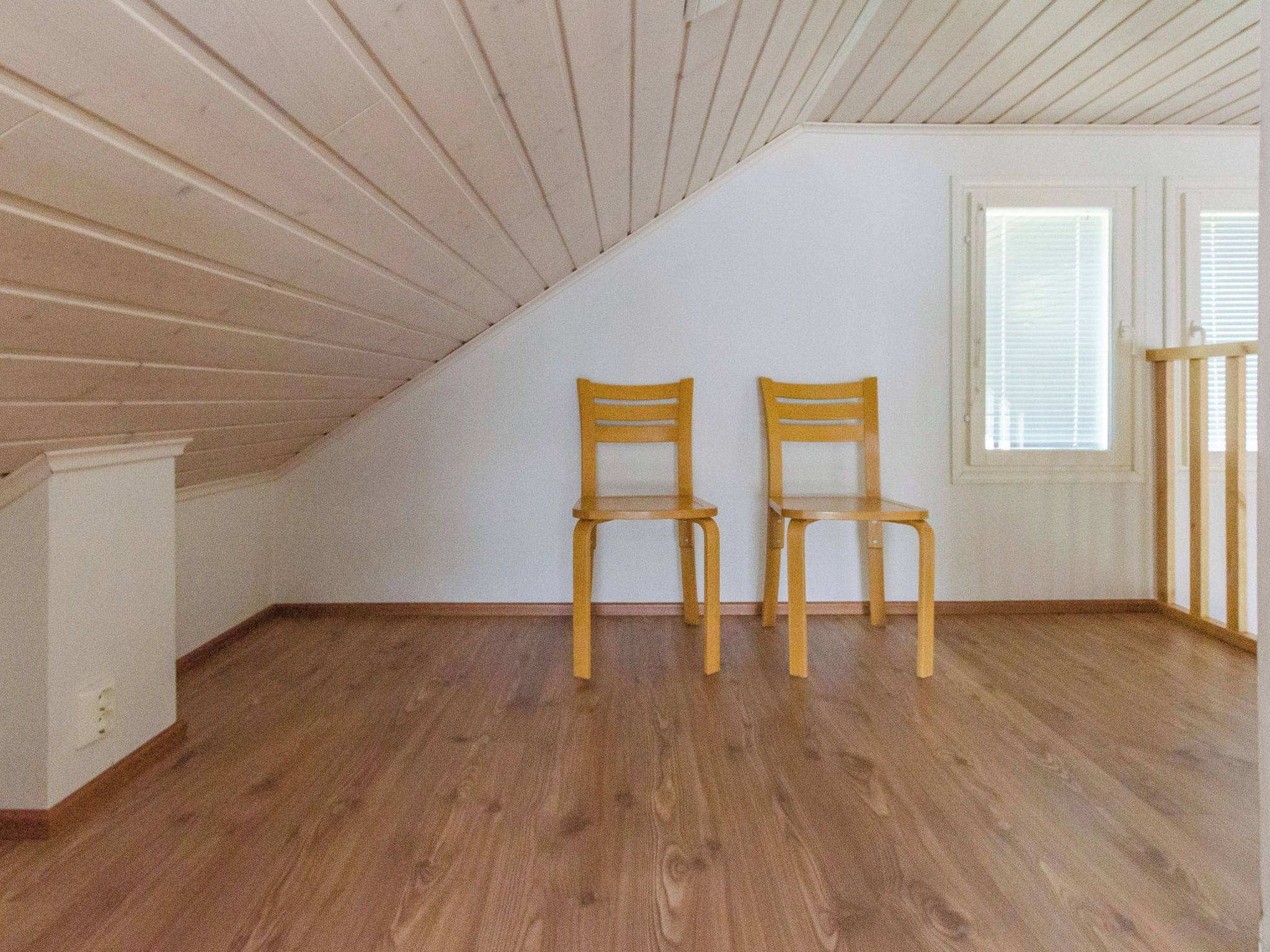 An attic room with wooden flooring, two wooden chairs, a window with blinds, and a white wall.