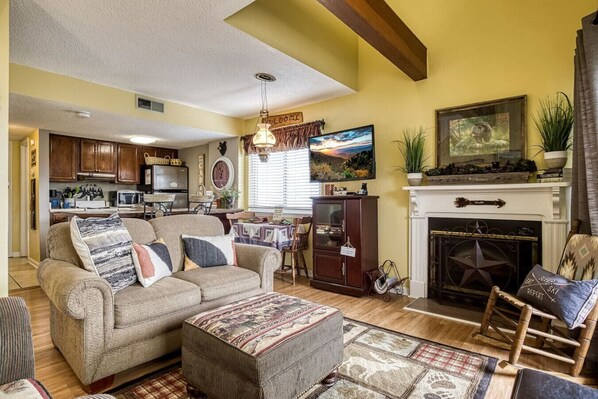 A cozy living room with a sofa, ottoman, and fireplace. There is a dining area with a table and chairs. The kitchen is visible in the background.
