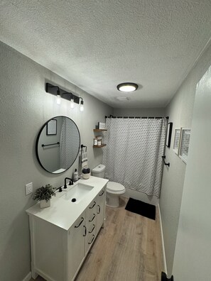 Een moderne badkamer met een wit wastafelmeubel, spiegel en toilet. Er is een douchegordijn met een patroon, een klein plantje op het wastafelmeubel en een zwarte mat op de vloer.