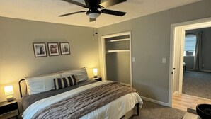 A bedroom with a bed, a ceiling fan, a nightstand, a lamp, framed pictures on the wall, and a wooden table with a pitcher. - Gig ‘Em Getaway (Santa Anna)