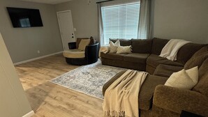 A living room with a ceiling fan, a large window with blinds, a brown sofa, a black armchair, and a patterned rug. - Gig ‘Em Getaway (Santa Anna)