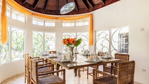 A dining area with a wooden table set for six, surrounded by wicker chairs, with large windows and curtains.