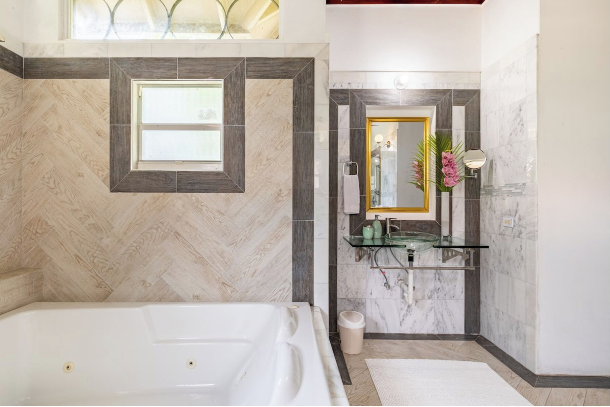 Un baño moderno con una bañera grande, un tocador con estante de vidrio y un espejo con marco dorado.