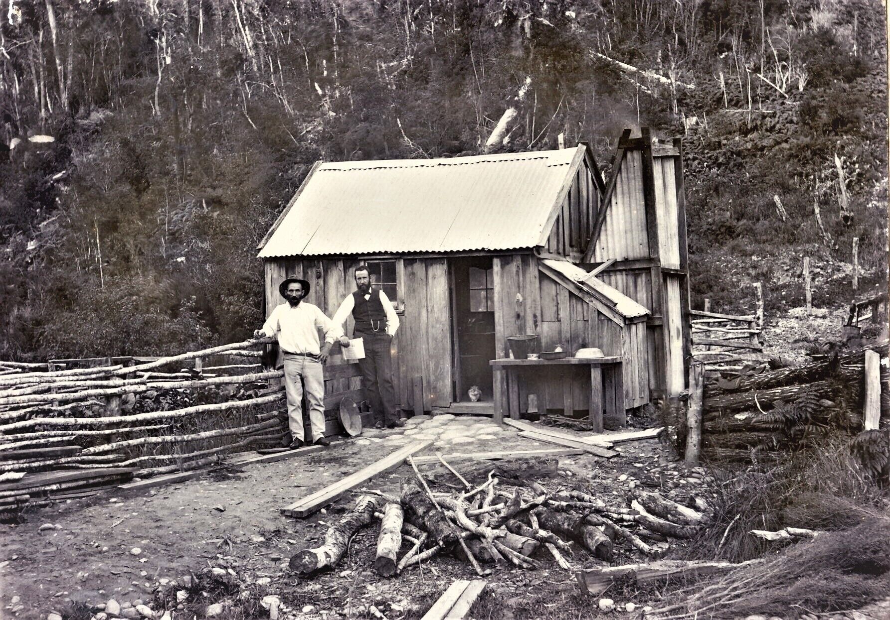 A rustic wooden cabin with a metal roof, surrounded by a wooden fence and a pile of logs.