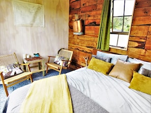 A cozy room with a bed, two chairs, a small table, and a map on the wall. - GOLD NUGGET HUT - Off-grid Glamping Site on a farm in Hokitika (Arahura Valley)