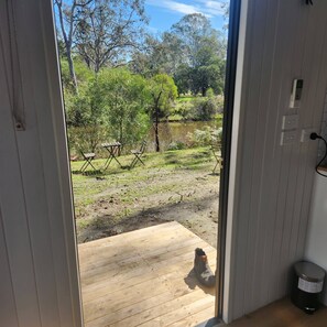 Blick vom Zimmerfenster auf ein Holzdeck, das in eine Grünanlage mit Bäumen und einem Gewässer führt.