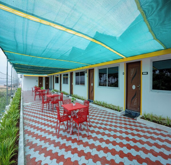 A covered outdoor seating area with red chairs and tables, a patterned floor, and a building with a wooden door and windows.