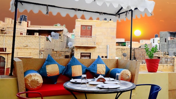 A rooftop terrace with a table set for two, a white canopy, and a view of the sunset over a cityscape. - Hotel Pleasure Haveli (Jaisalmer)