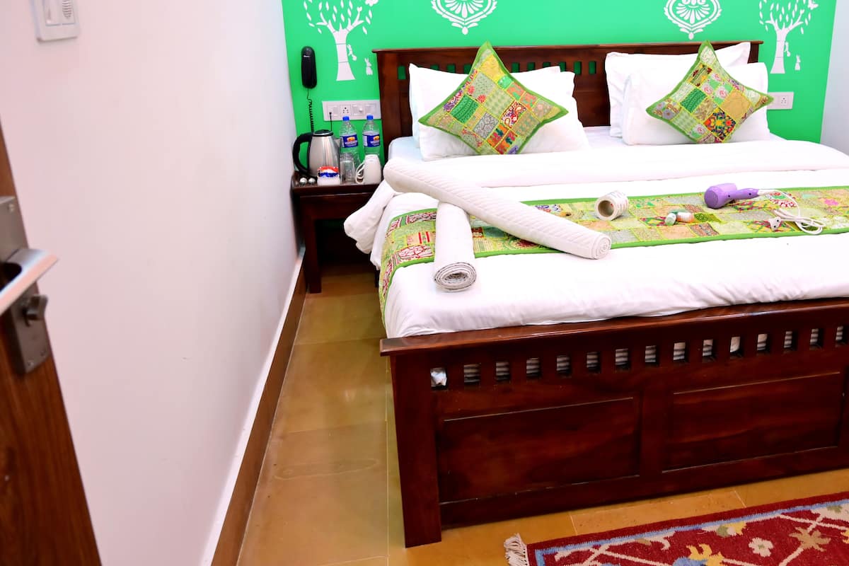 Ein ordentlich bezogenes Bett, eine grüne Wand mit weißen Bäumchen-Motiven, ein Nachttisch aus Holz mit einem Wasserkocher und Flaschen sowie ein Teppich mit Muster auf dem Boden.