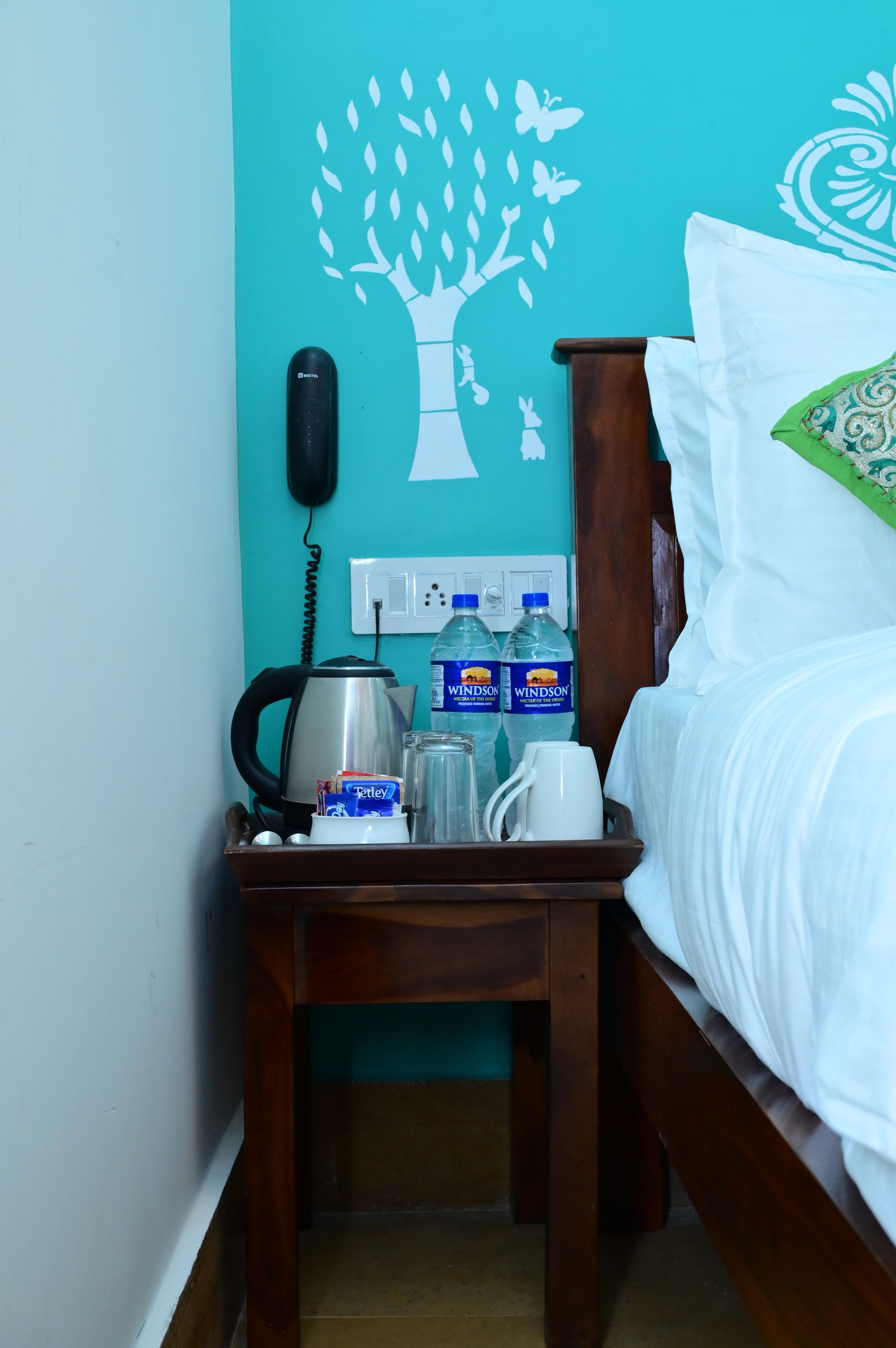 A neatly made bed with white bedding, a wooden nightstand with a kettle, a phone, and two water bottles.