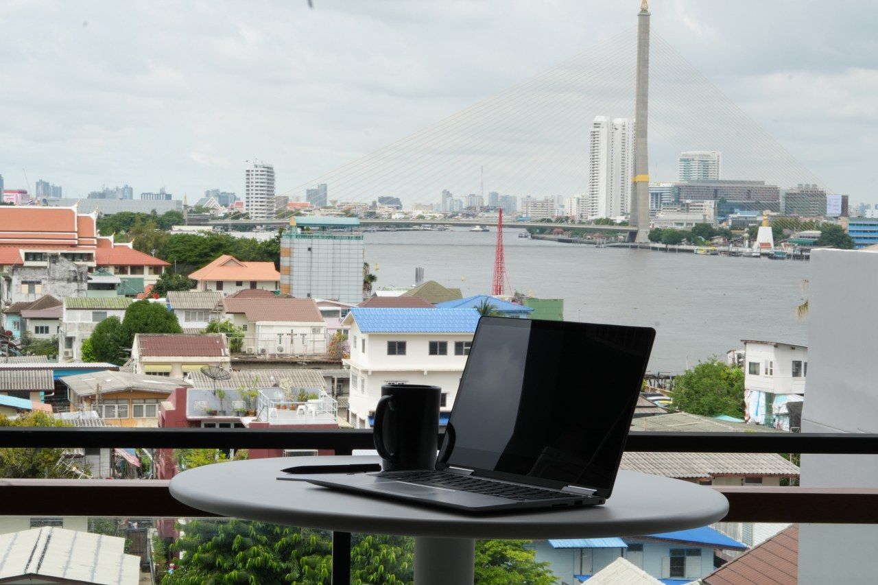A laptop on a balcony table with a view of a cityscape, including a river and a bridge.