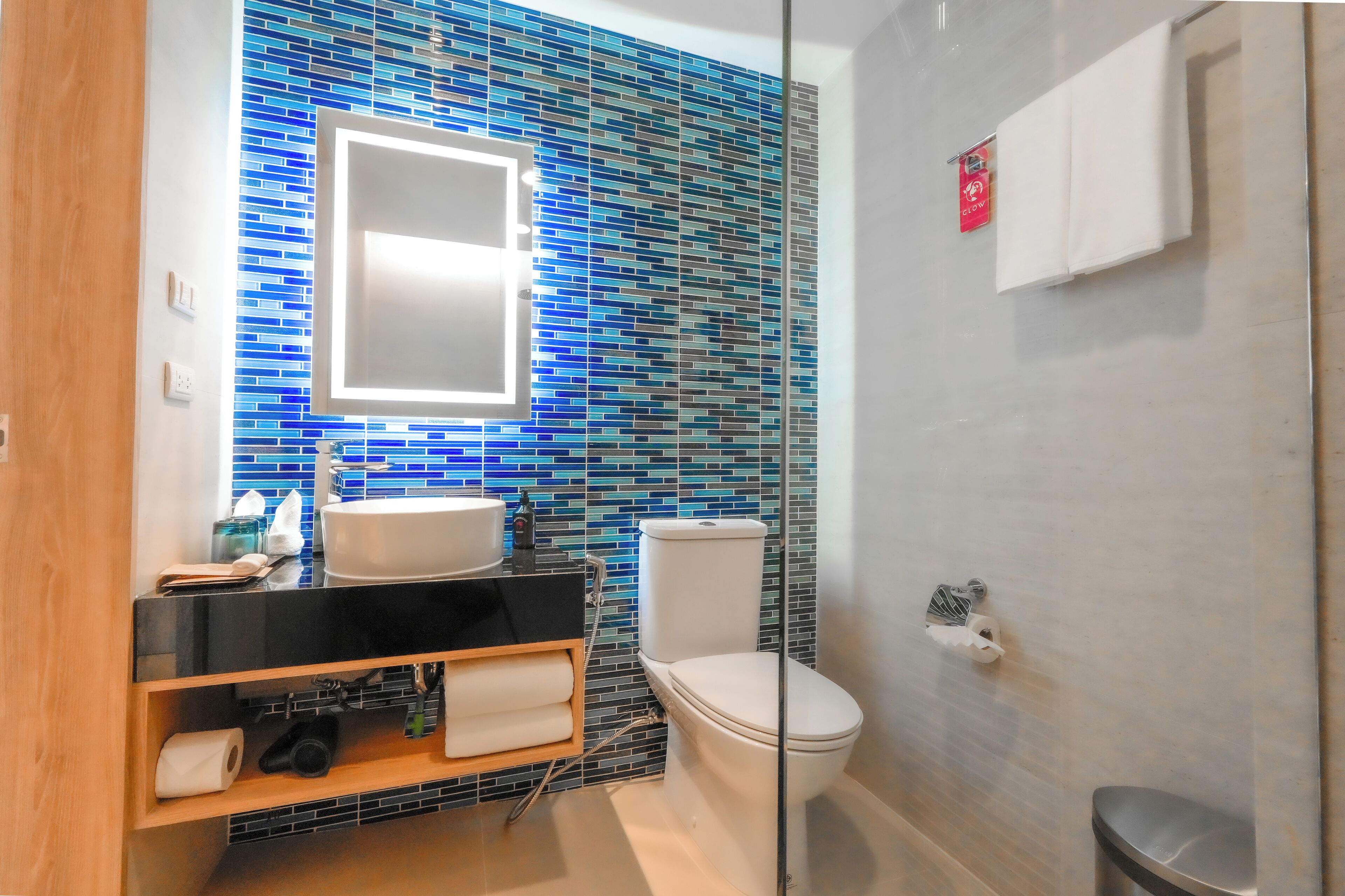A modern bathroom with a blue mosaic tile backsplash, a black countertop, a white sink, and a glass-enclosed shower.
