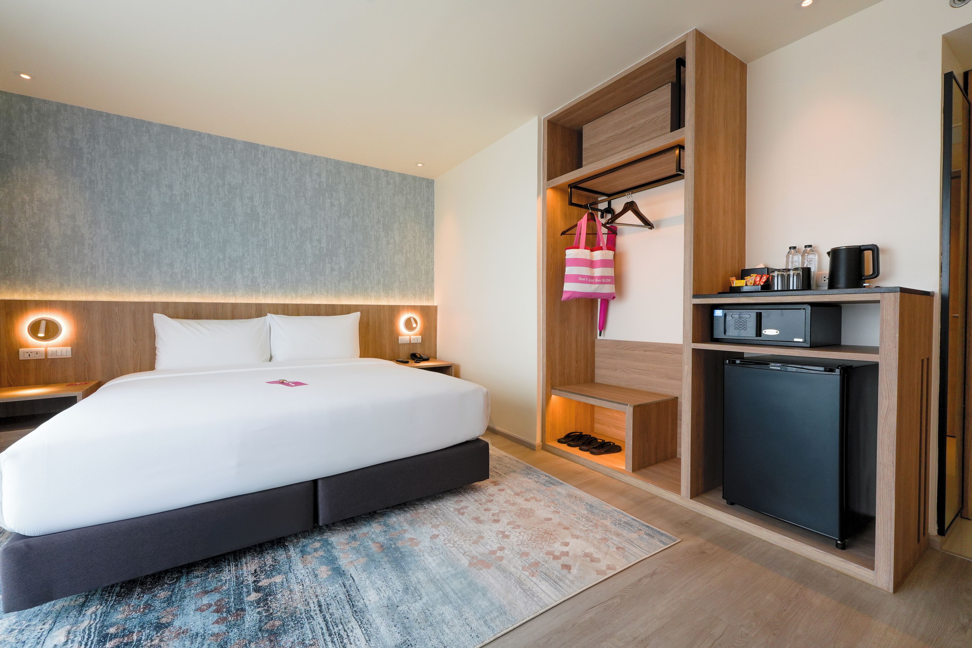 A modern hotel room with a large bed, bedside tables, a built-in mini-fridge, a microwave, and a hanging bag rack.