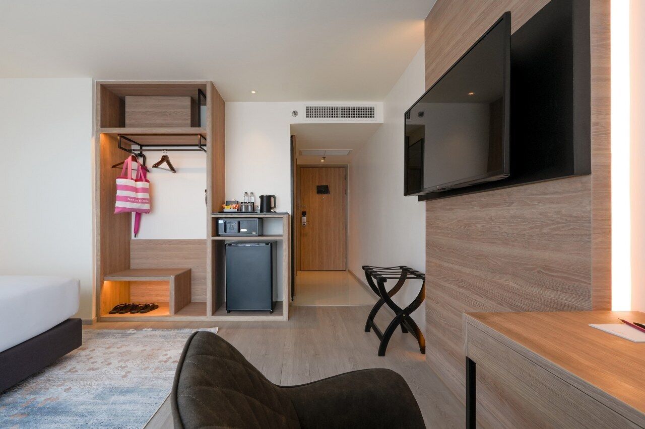 A modern hotel room with a bed, a desk, a TV, a mini-fridge, a small kitchenette, and a shoe rack.