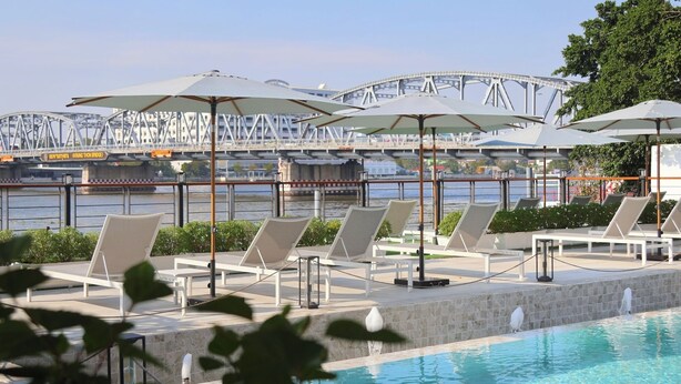 A poolside area with lounge chairs and umbrellas, a view of a bridge, and a body of water. - TARA Bangkok Riverside (Formerly GLOW Bangkok Riverside) (Bangkok)