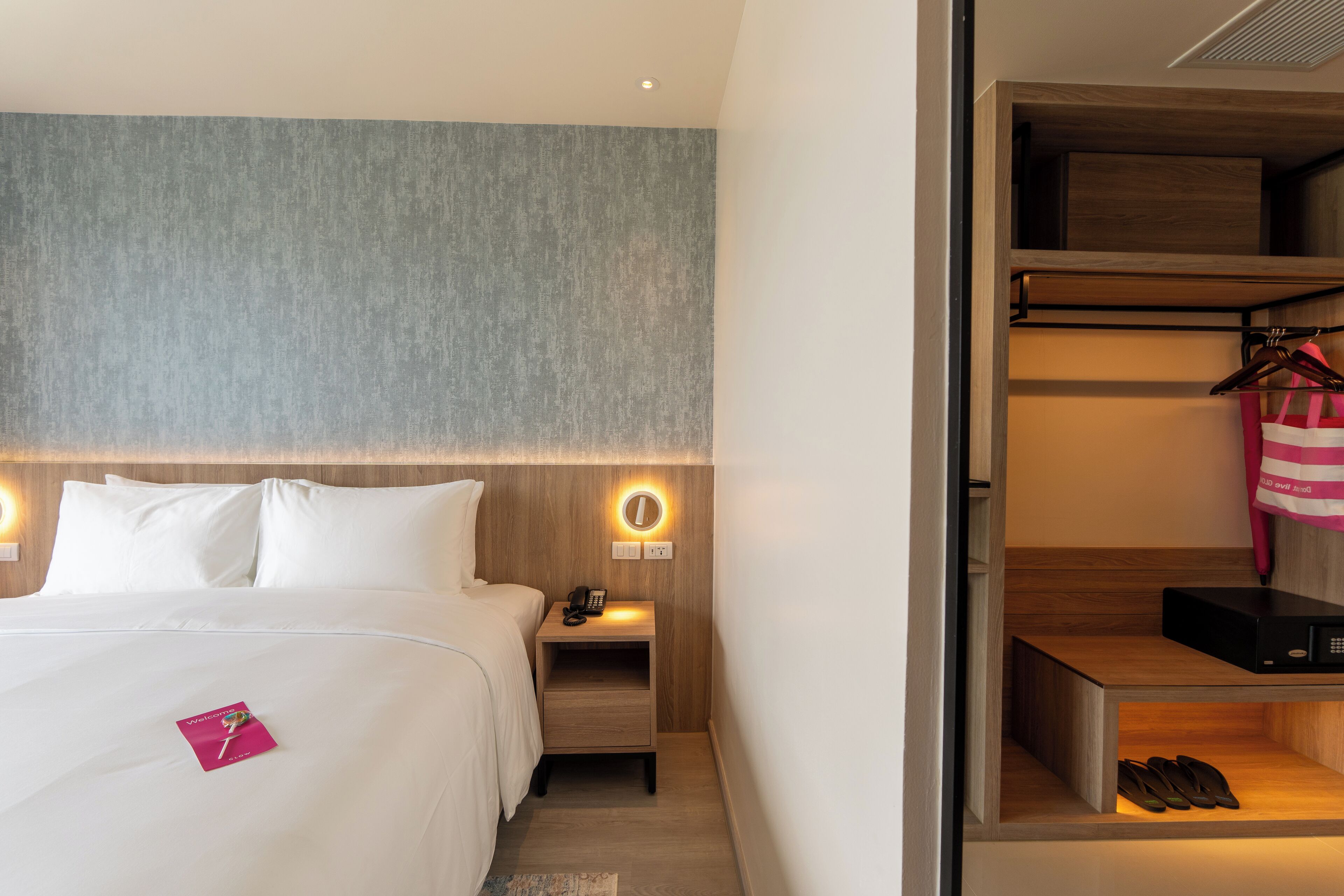 A hotel room with a neatly made bed, a bedside table with a lamp, a small shelf with shoes, and a hanging coat rack.