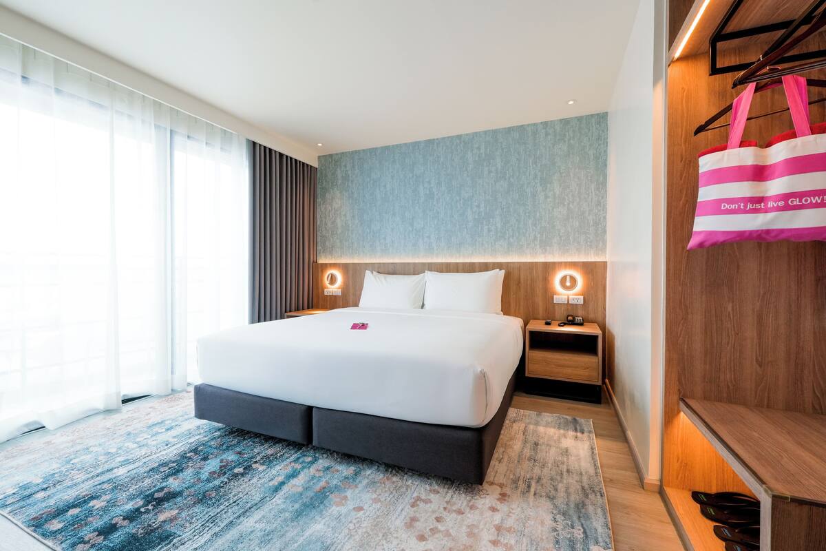 A modern hotel room with a large bed, bedside tables, a wooden wardrobe, and a large window with sheer curtains.