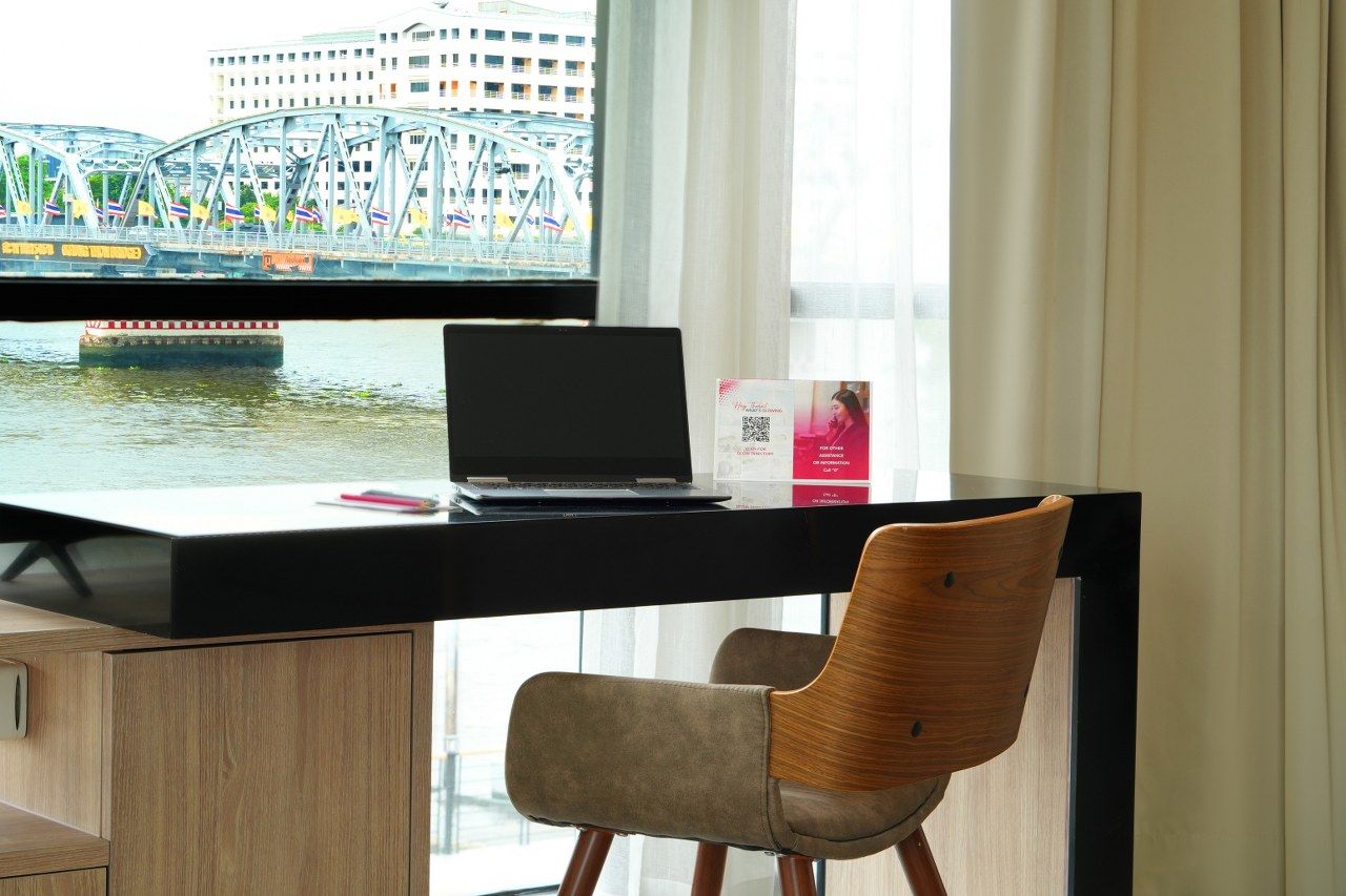 A modern hotel room with a desk, laptop, and chair, overlooking a river with a bridge and buildings.