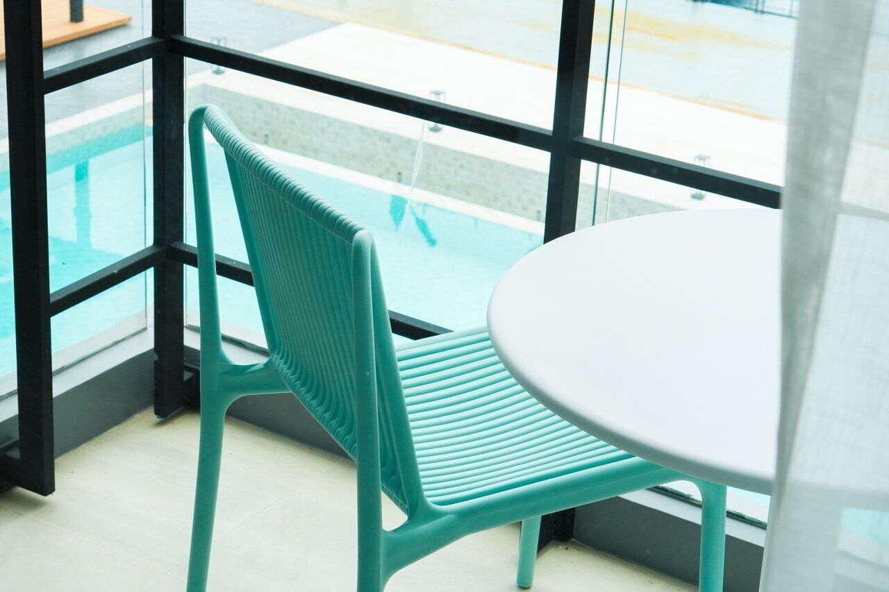 A corner of a room with a green chair and a white table, a window with a view of a pool, and a glass railing.